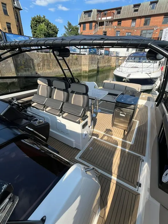 Luxury boat interior with grey seating, a table, and storage, set against a marina backdrop under a clear blue sky.